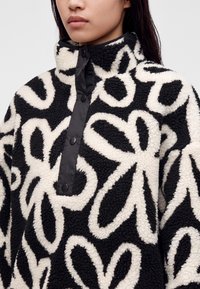 Femme portant un pull sherpa noir et blanc avec de grands motifs floraux et des boutons-pression sur la patte de col.