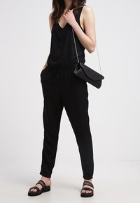 Czarny kombinezon z dekoltem w serek, elastyczną talią i bocznymi kieszeniami, w połączeniu z czarną, teksturowaną kopertówką i czarnymi płaskimi sandałami.