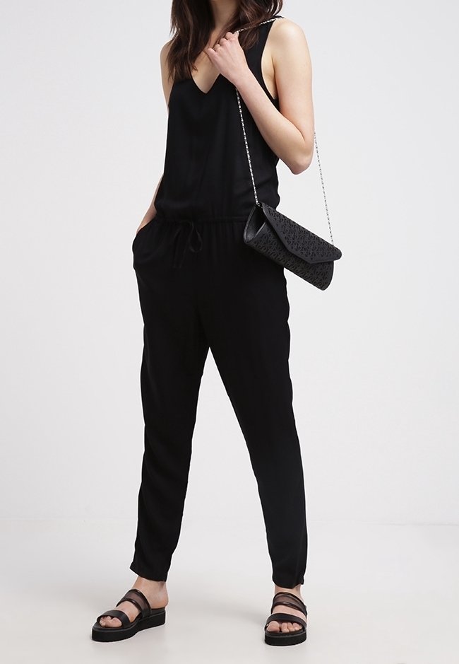 Czarny kombinezon z dekoltem w serek, elastyczną talią i bocznymi kieszeniami, w połączeniu z czarną, teksturowaną kopertówką i czarnymi płaskimi sandałami.
