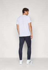 White short-sleeve t-shirt paired with dark navy athletic pants. Model wears white sneakers. The setting is plain and neutral in color.