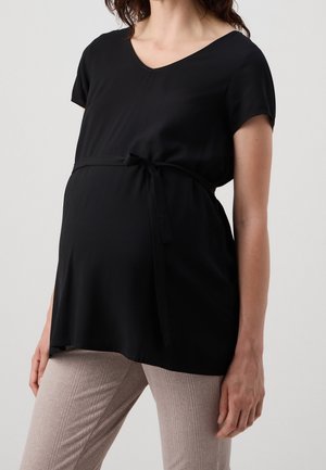 Těhotná žena v černém těhotenském topu s krátkým rukávem a vázáním v pase a béžových žebrovaných kalhotách na jednobarevném pozadí.