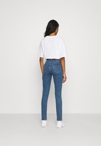 Jeans de tiro alto en azul con un ajuste slim, material de mezclilla, bolsillos traseros con detalles de costura, combinados con una camiseta blanca y zapatillas blancas.