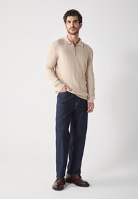 Polo beige à manches longues avec zip, associé à un jean en denim foncé et des chaussures marron. Le modèle se tient avec une main dans la poche.