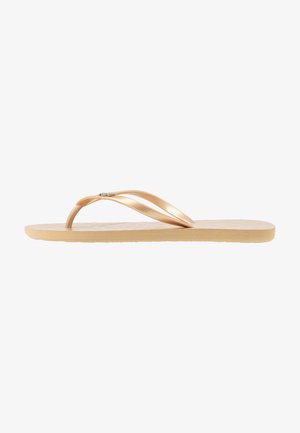 Sandal jepit dengan tali fleksibel berwarna emas dengan aksen dan sol beige bertexture. Desainnya datar dengan gaya minimalis.