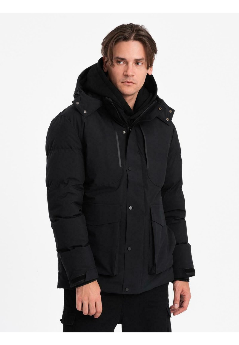 Giovane uomo che indossa un giubbotto invernale nero con cappuccio e grandi tasche a pattina, che guarda di lato su uno sfondo bianco semplice.