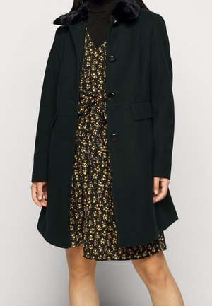 Femme portant un manteau noir avec un col en fourrure par-dessus une robe noire à petits motifs floraux jaunes et une ceinture nouée à la taille.