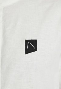 Tessuto bianco con finitura testurizzata, caratterizzato da una toppa nera. La toppa presenta un design astratto bianco ed è cucita sul materiale.