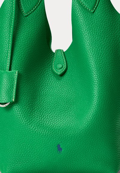 Zaļš teksturēts ādas rokassomiņa ar nelielu aizdari ar pogu un nemanāmu tumši zilu izšūtu logotipu netālu no pamatnes.