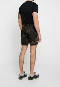 Man wearing black t-shirt, camo shorts, and black-white-yellow sneakers standing facing away against a plain white background.
