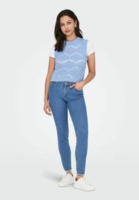 Chaleco de punto azul claro con un patrón hexagonal sobre una camiseta blanca, combinado con jeans de mezclilla azul ajustados y zapatillas blancas.