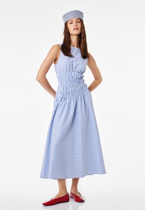 Femme portant une robe sans manches à carreaux bleus et blancs avec un chapeau assorti et des chaussures plates rouges, debout les mains sur les hanches.