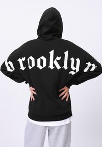 Hanorac negru cu un text mare alb "brooklyn" pe spate. Are un glugă cu șireturi și o croială relaxată. Purtați peste o cămașă albă și pantaloni gri.