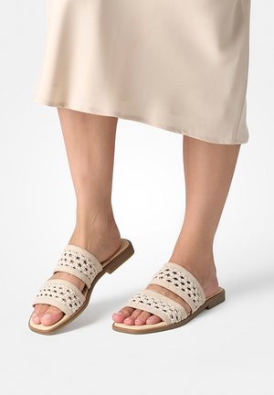 Unterschenkel und Füße in beigen Slip-on-Sandalen mit zwei gewebten Riemen und quadratischen Zehen, kombiniert mit einem hellbeigen Rock.