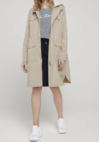 Parka beis resistente al agua con capucha ajustable, dos bolsillos frontales, cierre de cremallera, llevada sobre una camiseta a rayas, combinada con pantalones cortos azul marino y zapatillas blancas.