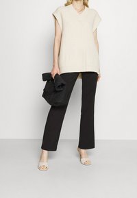 Beige gebreide mouwloze top met een V-hals, gecombineerd met zwarte broek en lichtbeige open-toe mules, een zwarte gewatteerde tas houdend.