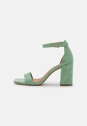 Zielone sandały na obcasie z ekoskóry o fakturze krokodyla, z odkrytymi palcami i regulowanym paskiem na kostce. Kwadratowy nosek i masywny obcas.