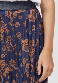 Marinblå plisserad kjol med brunt blommönster. Har en texturerad svart midja och slät tyg med vertikala ränder. Hand visas.