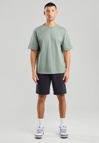 Green cotton t-shirt with short sleeves, paired with dark shorts. Man standing in white sneakers, plain background.