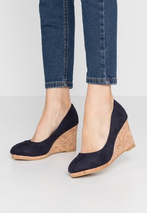 Námořnické modré semišové klínové boty s korekovou podešví a zaobleným špičkou, umístěné na denimu a světlem pozadí.