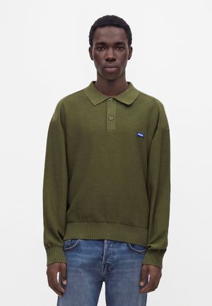 Joven con pelo corto que lleva una camisa polo de manga larga color verde oliva y vaqueros azules, de pie frente a un fondo claro y liso.