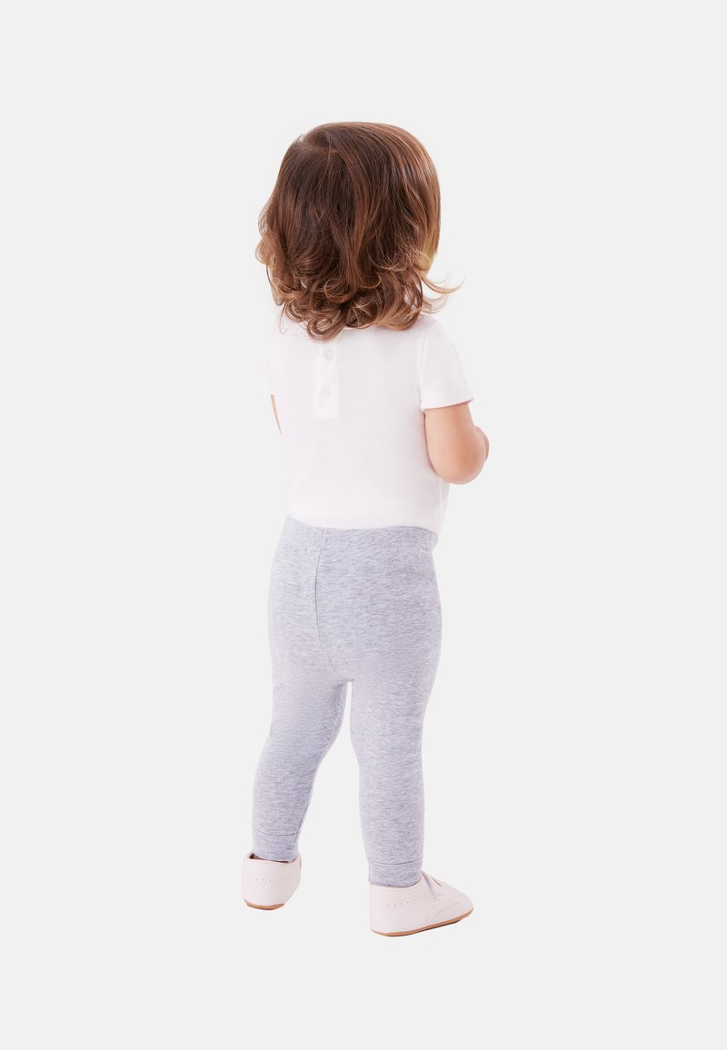 Kleinkind mit lockigem braunem Haar, das ein weißes Hemd, graue Leggings und hellrosa Schuhe trägt und mit dem Rücken zum Betrachter vor einem weißen Hintergrund steht.