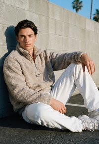 Fiatal férfi ül az úttesten, egy betonfalnak támaszkodva, bézs polárkabátot, fehér nadrágot és tornacipőt visel, napsütésben.