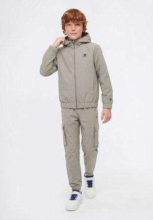 Jeune garçon aux cheveux roux portant une veste à capuche grise et un pantalon cargo, ajustant le col de la veste, debout devant un fond blanc uni.