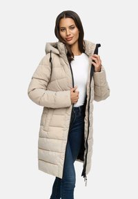 Vrouw in een beige lange gewatteerde jas met capuchon, witte shirt en blauwe jeans, die de jas met één hand bij de kraag openhoudt.