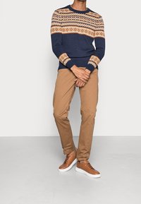 Pull bleu marine avec des accents beige et rouille à motifs, poignets côtelés, assorti à un pantalon chino beige et des baskets marron à semelles blanches.