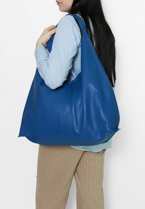 Vrouw met lang donker haar draagt een grote blauwe leren schoudertas, draagt een lichtblauw overhemd en beige broek met fijne strepen, zijaanzicht.