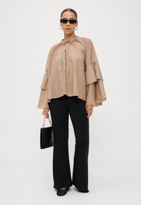 Beige, lightweight blouse with a drawstring neck, layered bell sleeves, paired with black trousers and black flat shoes, holding a black purse.