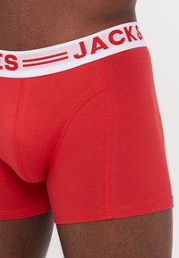 Rote Baumwoll-Boxershorts mit einem weißen Elastikbund, der "JACK&JONES" in roter Schrift zeigt. Zwei Seitennähte, glatte Textur.