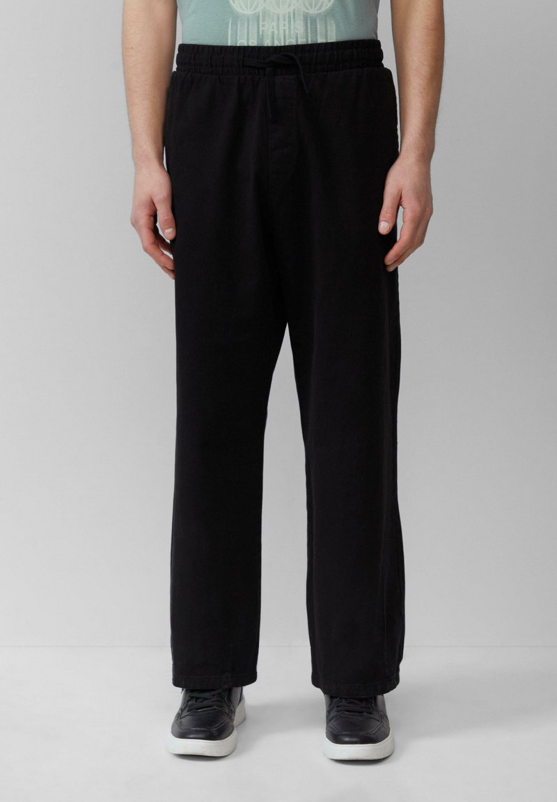 Schwarze, lässig geschnittene Hose mit elastischem Bund und Kordelzug; glattes Material und gerade Beine; kombiniert mit schwarzen Sneakern.