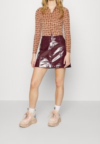 Hnědý vzorovaný top s dlouhým rukávem a límcem, v kombinaci s burgundy lesklou sukní ve tvaru A se zapínáním na knoflíky vpředu. Celý outfit doplňují světlé tenisky.