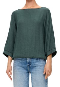 Blusa verde oscuro con un acabado texturizado, escote amplio de barco y mangas tres cuartos. Combinada con vaqueros azul claro.