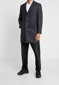 Grå uldfrakke med en skræddersyet pasform, tre knapper og sidelommer, parret med sorte denimbukser og sorte sportsko.