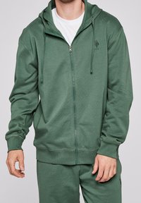Uomo che indossa una felpa verde con zip e cappuccio con cordini e pantaloni della tuta verdi abbinati su una maglietta bianca, davanti a uno sfondo semplice.