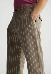 Olivgrüne gestreifte Hose mit pinken Streifen, flaches Vorderteil, seitliche Taschen und Gürtelschlaufen, aus einem strukturierten Stoff gefertigt.