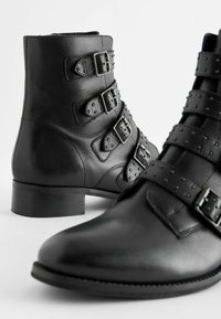 Czarne skórzane botki za kostkę z wieloma ćwiekowanymi paskami, srebrnymi klamrami, okrągłym noskiem i niskim, grubym obcasem. Gładka tekstura.