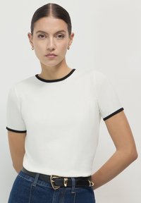 Top bianco a maniche corte con bordi neri. Design aderente e texture liscia. Abbinato a jeans blu a vita alta e una cintura nera con accessori dorati.