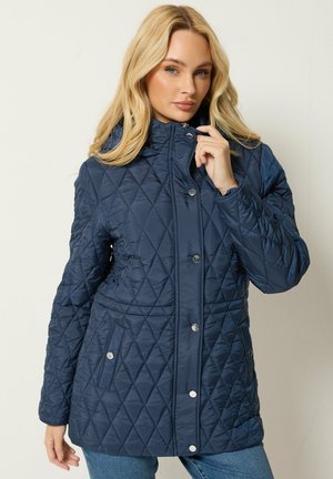 Femme blonde portant une veste matelassée bleu marine avec des boutons-pression, debout devant un fond clair uni.