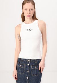 Bela rebrasta majica na naramenice z osrednjim črnim logotipom "CK" in besedilom "Calvin Klein Jeans", v kombinaciji z denim krilom z gumbi.