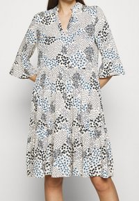 Kukallinen mekko, jossa on beige pohja ja sinisiä sekä mustia kukkakuosisia kuvioita, V-pääntie ja kolmannesihdattu liehuva hiha.