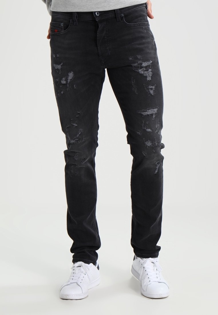 Hombre vistiendo jeans ajustados negros desgastados y zapatillas blancas, de pie con las manos cerca de los bolsillos frente a un fondo liso.