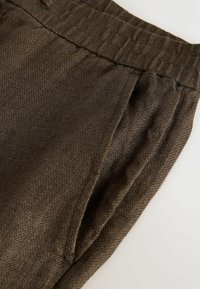 Détail d’un pantalon en tissu marron texturé avec taille élastique et poche avant visible sur un fond clair.