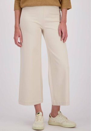 Vrouw in beige wijde cropped broek, een bruine trui en crèmesneakers met luipaardprintdetail op de hiel.