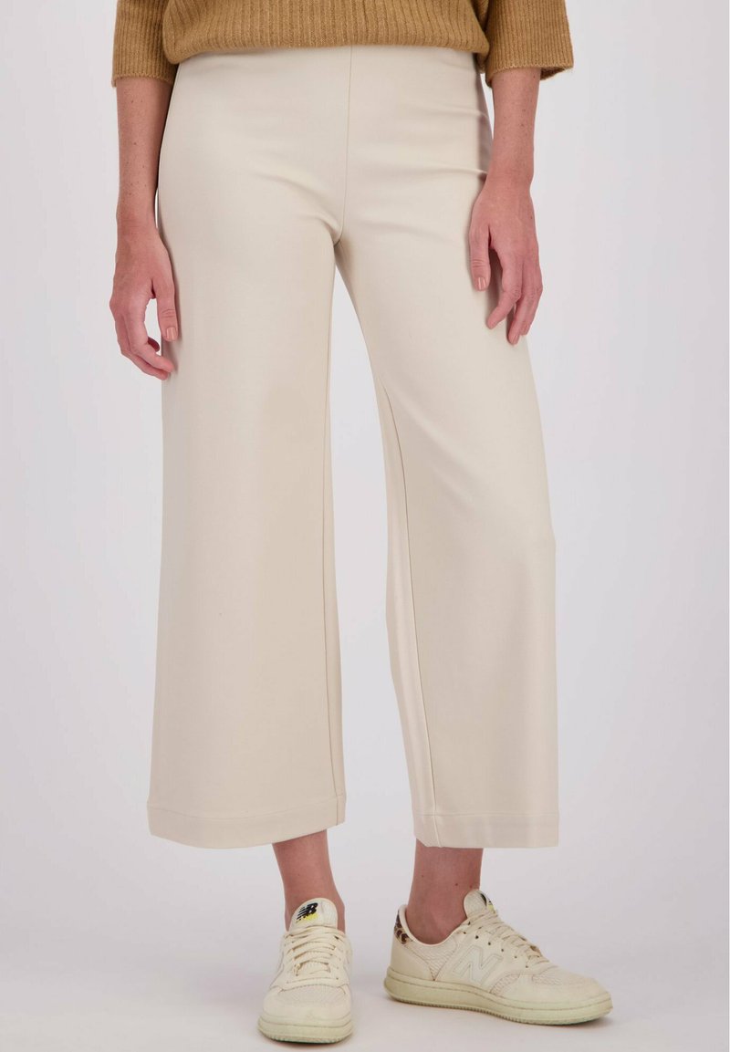 Vrouw in beige wijde cropped broek, een bruine trui en crèmesneakers met luipaardprintdetail op de hiel.