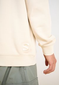 Kremowy bawełniany sweatshirt z ribbowanymi mankietami i dołem, z haftowanym logo "S" po stronie dolnej. Materiał ma gładką teksturę.