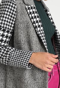 Ein schwarz-weißer Hahnentritt-Blazer mit einem Fischgrätenmuster, ausgestattet mit einem Schalkragen und kontrastierend strukturierten Ärmeln.