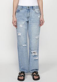 Svetlo modri jeans s sproščenim krojem, ki imajo obrabljena mesta in razfrčan rob. V kombinaciji s črnimi sandali, ki prikazujejo dekorativne dodatke.